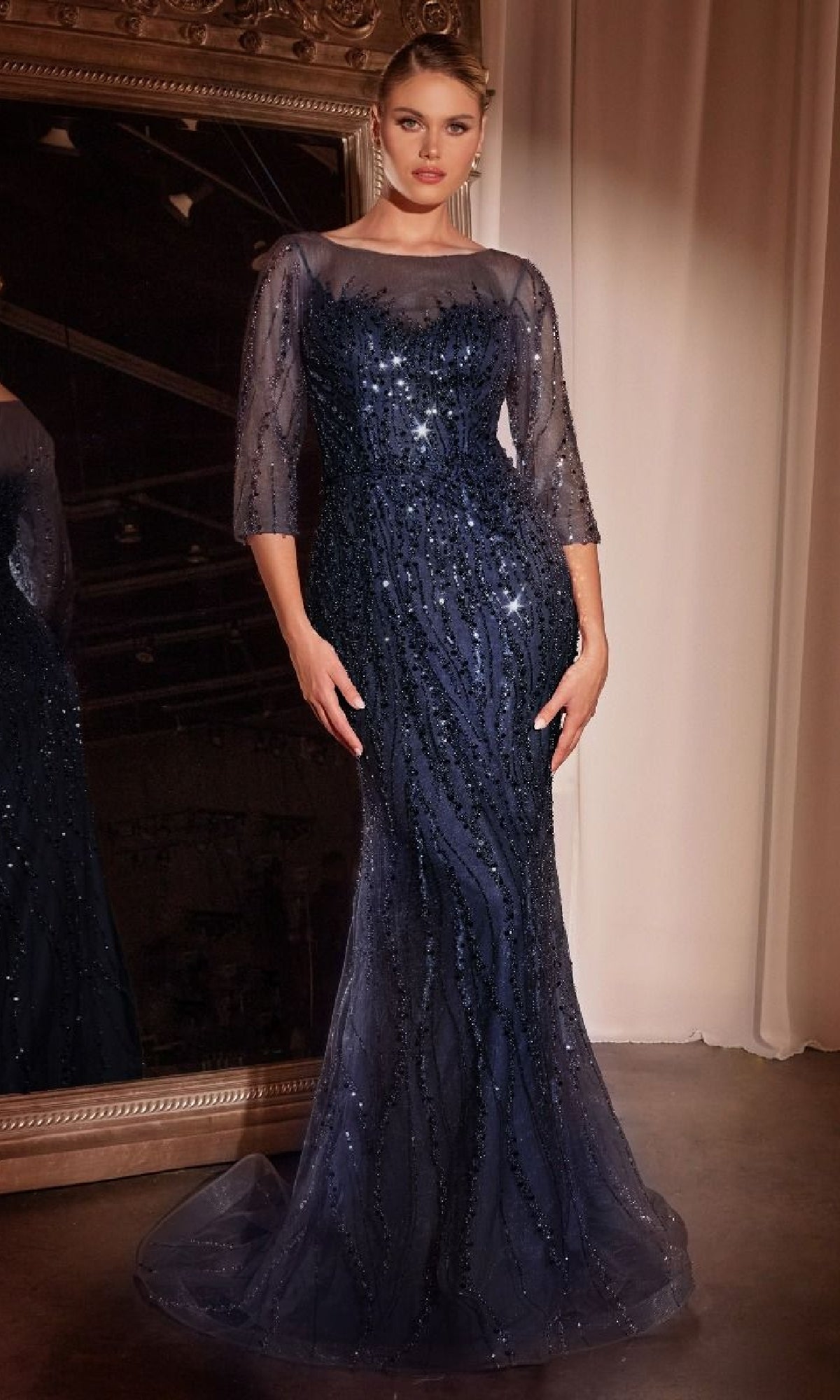 A woman stands before a mirror wearing the Ladivine CD0105, a dark blue long sleeve sequin prom gown with sheer sleeves, sparkling embellishments, and an elegant mermaid skirt.